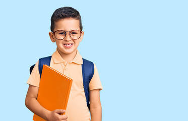 Obraz premium Little cute boy kid wearing school bag and holding book looking positive and happy standing and smiling with a confident smile showing teeth