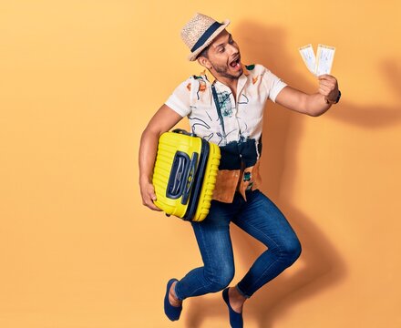 Young handsome latin man on vacation wearing summer clothes smiling happy. Jumping with smile on face holding cabin bag and airplane boarding pass over isolated background