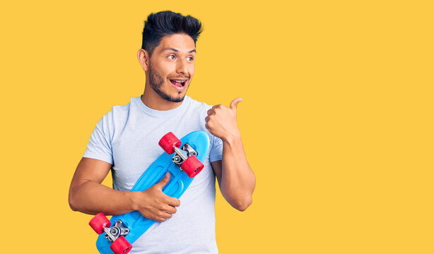 Handsome latin american young man holding skate pointing thumb up to the side smiling happy with open mouth
