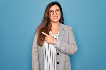 Young hispanic business woman wearing glasses standing over blue isolated background cheerful with a smile on face pointing with hand and finger up to the side with happy and natural expression