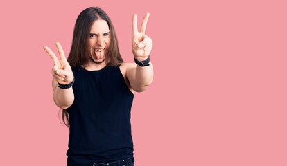 Young adult man with long hair wearing goth style with black clothes smiling with tongue out...