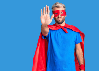 Naklejka premium Young blond man wearing super hero custome doing stop sing with palm of the hand. warning expression with negative and serious gesture on the face.
