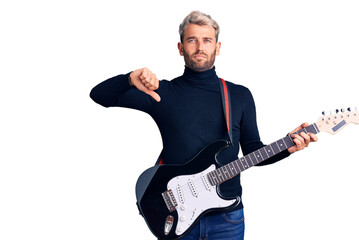 Young handsome blond man playing electric guitar with angry face, negative sign showing dislike with thumbs down, rejection concept