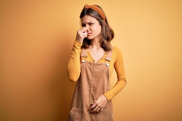 Young beautiful blonde girl wearing overall standing over yellow isolated background smelling...