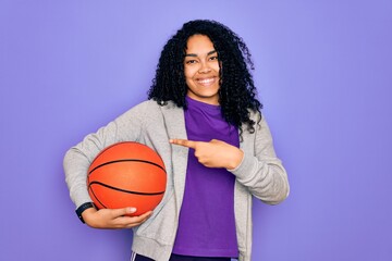 African american curly sportswoman doing sport holding basketball ball over purple background very happy pointing with hand and finger