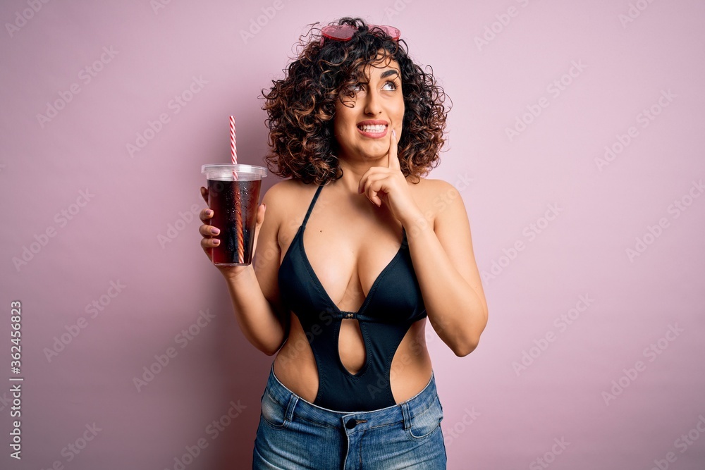 Poster beautiful arab woman on vacation wearing swimsuit drinking cola refreshment using straw serious face