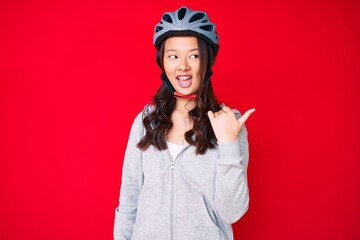 Young beautiful chinese girl wearing bike helmet smiling with happy face looking and pointing to the side with thumb up.