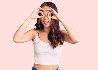 Young beautiful chinese girl wearing casual clothes doing ok gesture like binoculars sticking tongue out, eyes looking through fingers. crazy expression.