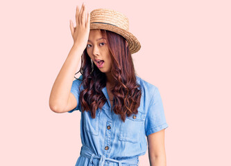 Young beautiful chinese girl wearing summer hat surprised with hand on head for mistake, remember error. forgot, bad memory concept.