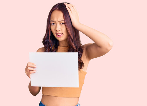 Young beautiful chinese girl holding paper banner with blank space stressed and frustrated with hand on head, surprised and angry face