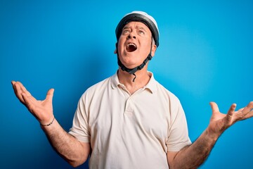 Naklejka premium Middle age hoary cyclist man wearing bike security helmet over isolated blue background crazy and mad shouting and yelling with aggressive expression and arms raised. Frustration concept.
