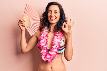 Young beautiful hispanic woman wearing bikini and hawaiian lei holding hand fan doing ok sign with...