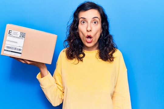 Young beautiful hispanic woman holding delivery package scared and amazed with open mouth for surprise, disbelief face