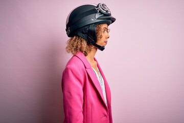 African american motorcyclist woman with curly hair wearing moto helmet over pink background looking to side, relax profile pose with natural face with confident smile.