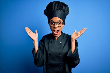 Young african american chef woman wearing cooker uniform and hat over blue background celebrating crazy and amazed for success with arms raised and open eyes screaming excited. Winner concept