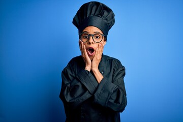 Young african american chef woman wearing cooker uniform and hat over blue background afraid and shocked, surprise and amazed expression with hands on face