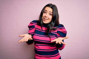 Young brunette elegant woman wearing striped shirt over pink isolated background clueless and confused expression with arms and hands raised. Doubt concept.