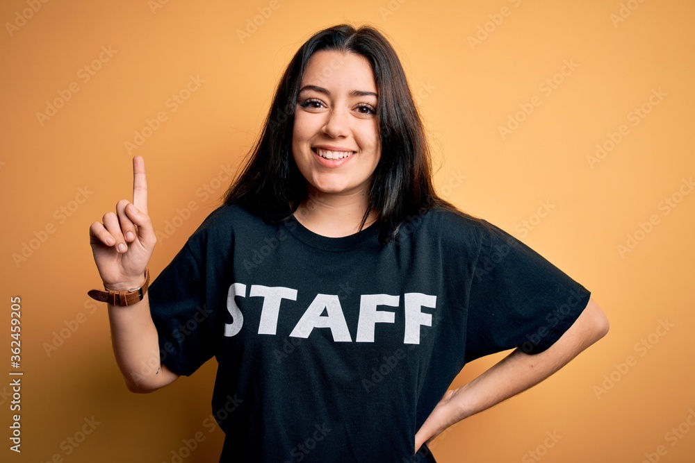 Wall mural young brunette worker woman wearing staff t-shirt as uniform over yellow isolated background surpris