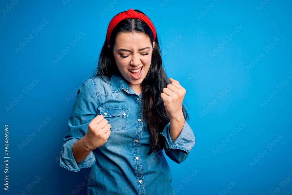 Wall mural Young brunette woman wearing casual denim shirt over blue isolated background celebrating surprised and amazed for success with arms raised and eyes closed. Winner concept.