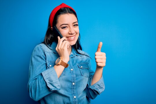 Young brunette woman talking on smartphone over blue isolated background happy with big smile doing ok sign, thumb up with fingers, excellent sign