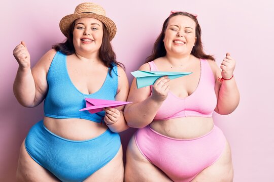 Young plus size twins wearing bikini holding paper airplane screaming proud, celebrating victory and success very excited with raised arm