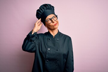 Middle age brunette chef woman wearing cooker uniform and hat over isolated pink background confuse and wondering about question. Uncertain with doubt, thinking with hand on head. Pensive concept.