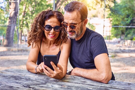 Middle age couple smiling happy using smartphone. Sitting on the bench at park