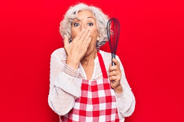 Senior grey-haired woman wearing apron holding whisk covering mouth with hand, shocked and afraid...