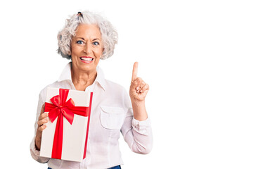 Senior grey-haired woman holding gift surprised with an idea or question pointing finger with happy face, number one