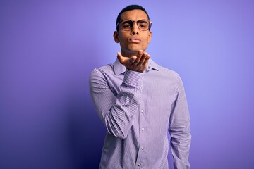 Handsome african american man wearing striped shirt and glasses over purple background looking at the camera blowing a kiss with hand on air being lovely and sexy. Love expression.
