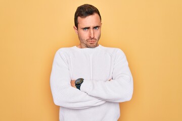 Young handsome man with blue eyes wearing casual sweater standing over yellow background skeptic and nervous, disapproving expression on face with crossed arms. Negative person.