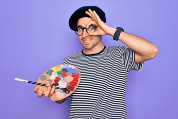 Young artist man with blue eyes wearing french beret and glasses drawing using paintbrush with happy face smiling doing ok sign with hand on eye looking through fingers