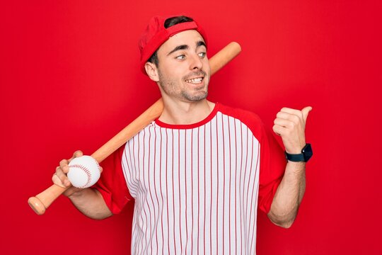 Young handsome sporty man with blue eyes playing baseball wearing cap holding bat and ball pointing and showing with thumb up to the side with happy face smiling