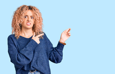 Young blonde woman with curly hair wearing casual winter sweater pointing aside worried and nervous...