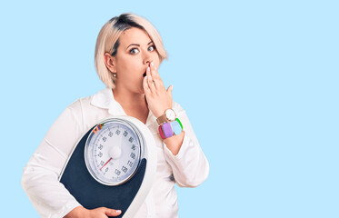 Young blonde plus size woman holding weighing machine covering mouth with hand, shocked and afraid...