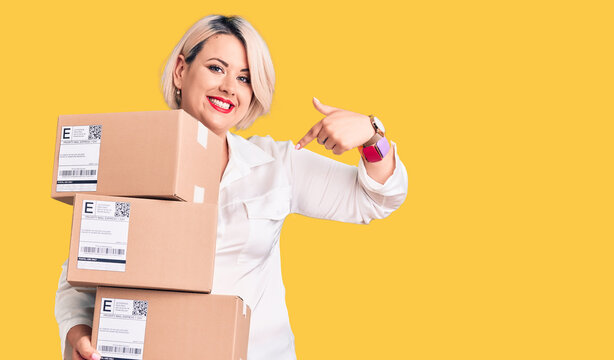 Young blonde plus size woman holding delivery cardboard package pointing finger to one self smiling happy and proud