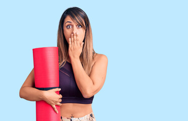 Young beautiful woman wearing sportswear holding yoga mat covering mouth with hand, shocked and...