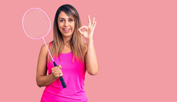 Young beautiful woman holding badminton racket doing ok sign with fingers, smiling friendly gesturing excellent symbol