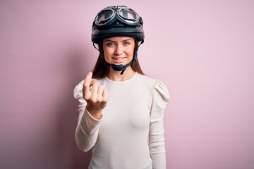 Young beautiful motorcyclist woman with blue eyes wearing moto helmet over pink background Beckoning come here gesture with hand inviting welcoming happy and smiling