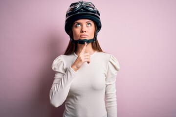Young beautiful motorcyclist woman with blue eyes wearing moto helmet over pink background Thinking concentrated about doubt with finger on chin and looking up wondering