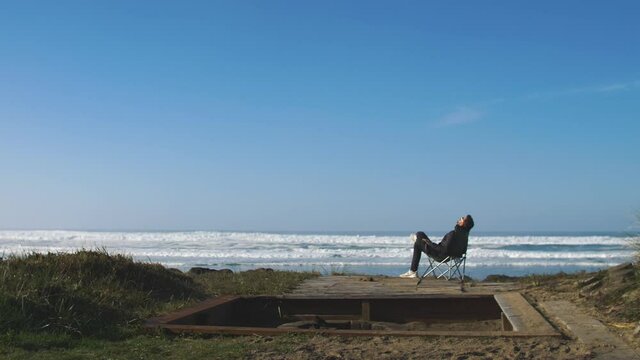 Man sitting in folding chair at the beach and watches the waves in the ocean and thinking about life - relaxing holiday - freedom - alone and+d introverted, 4k footage slow motion