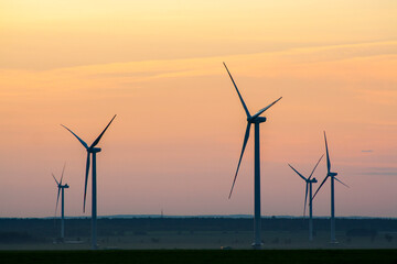 Fototapeta premium many windmills stand on a large field spinning in the evening against a sunset orange sky. Alternative Energy Concept