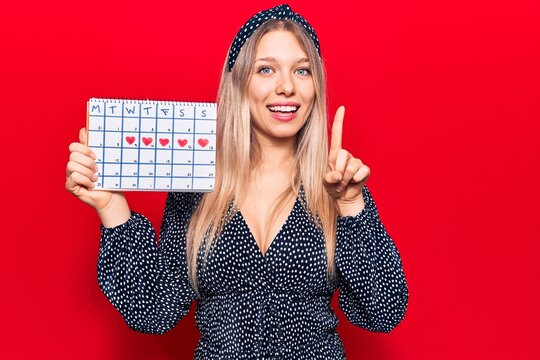 Young blonde woman holding heart calendar smiling with an idea or question pointing finger with happy face, number one