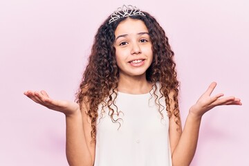 Beautiful kid girl with curly hair wearing princess tiara clueless and confused with open arms, no...