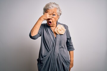 Senior beautiful grey-haired woman wearing casual dress standing over white background peeking in shock covering face and eyes with hand, looking through fingers with embarrassed expression.