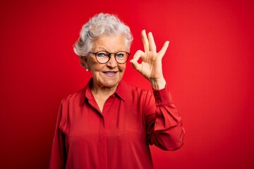Senior beautiful grey-haired woman wearing casual shirt and glasses over red background smiling...