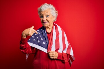 Senior beautiful grey-haired patriotic woman wearing united states flag over red background with surprise face pointing finger to himself