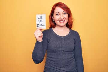 Young beautiful redhead woman asking for equality holding paper with we are equal message looking positive and happy standing and smiling with a confident smile showing teeth