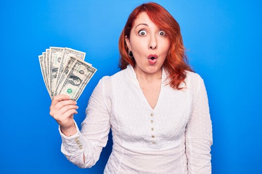 Young beautiful redhead woman holding bunch of dollars banknotes over blue background scared and amazed with open mouth for surprise, disbelief face