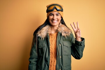 Young beautiful hispanic woman wearing ski glasses and coat for winter weather showing and pointing up with fingers number three while smiling confident and happy.